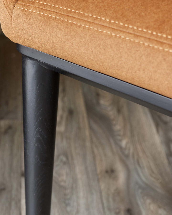 Close-up of a chair, showing a brown upholstered seat with stitch detailing and a black wooden leg.