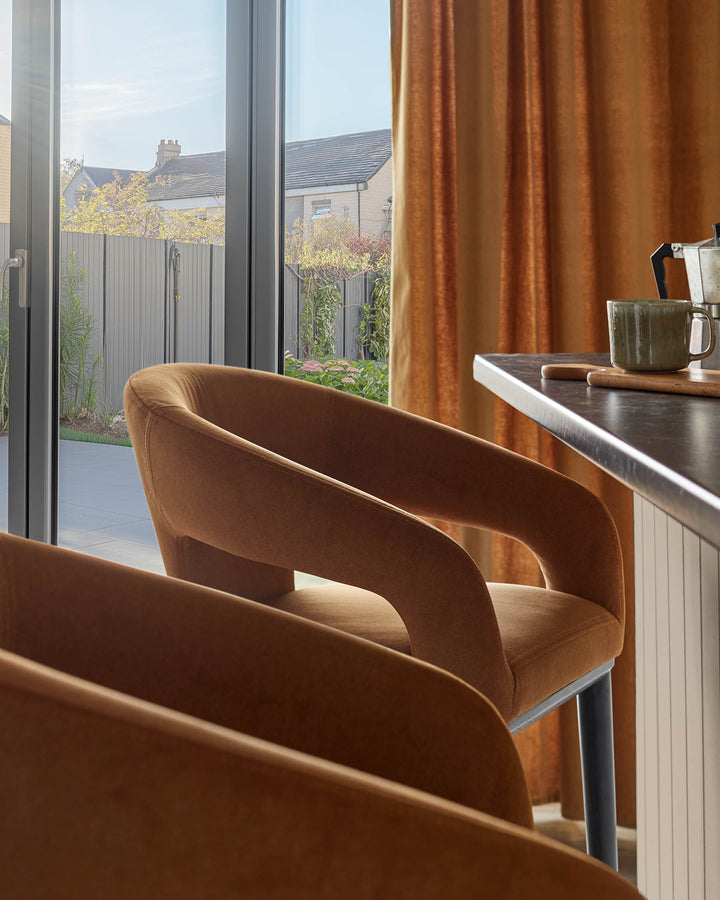 Modern brown chairs at a kitchen island with dark worktop and copper curtains. Outside is a garden visible through doors.