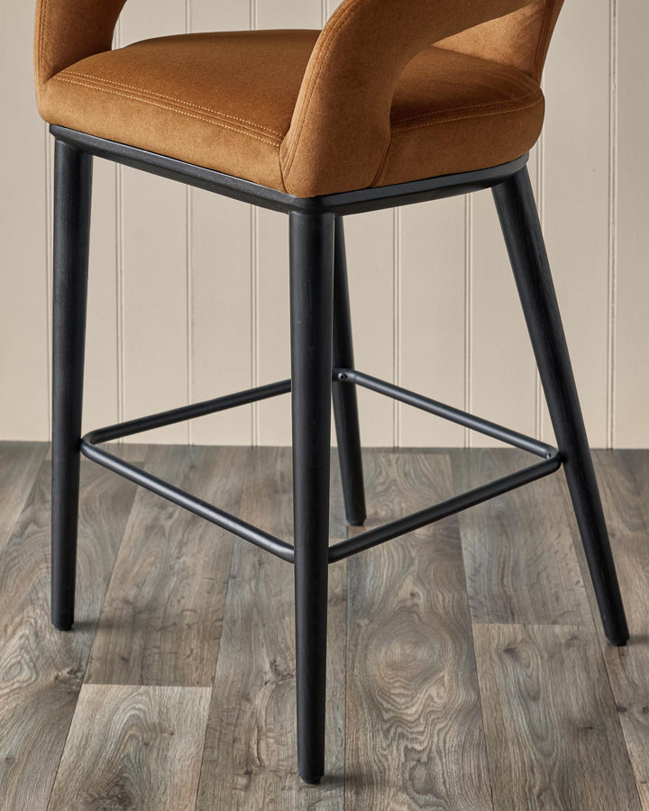 A stool with a brown seat and black legs, with a supportive footrest, on a wooden floor.