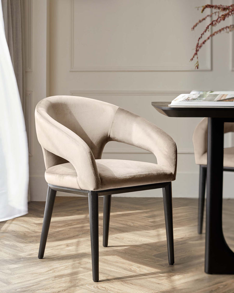 Elegant contemporary beige velvet dining chair with a curved backrest and cushioned seat, featuring dark wooden legs, beside a round black dining table with a magazine on top, set against a neutral wall and light wood flooring.