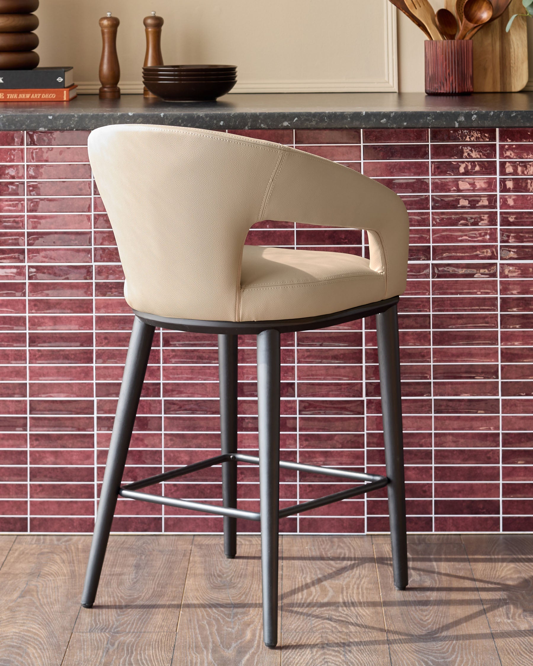 A stylish, light beige bar stool with black legs sits against a red tiled splashback in a modern kitchen setting.