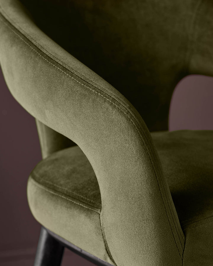 Close-up of a stylish, olive-green velvet chair with a curved armrest and dark wooden legs, exuding luxury and comfort.