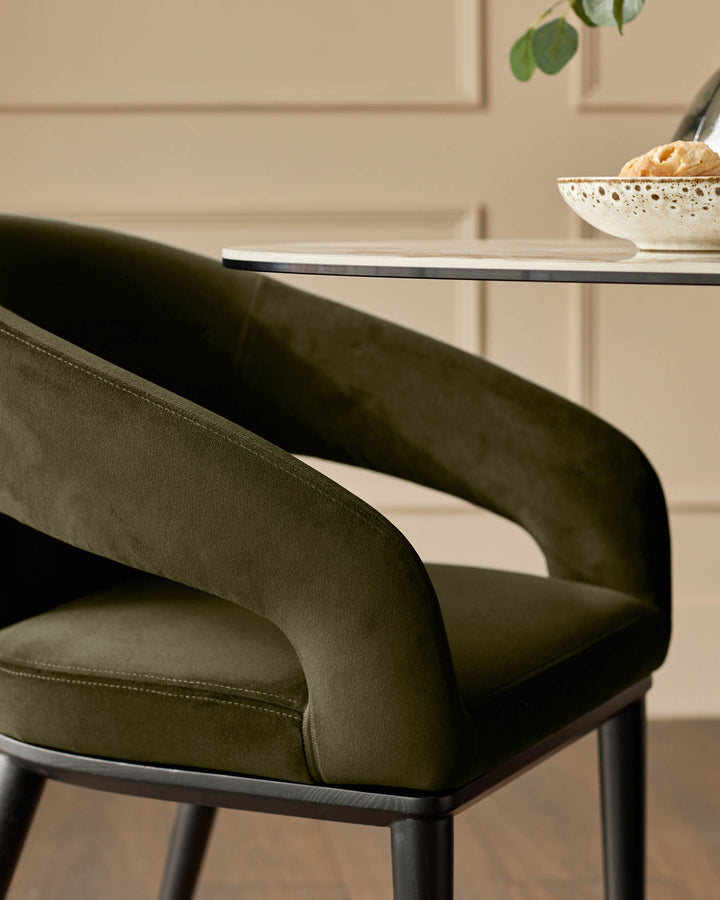 Close-up of a stylish, olive green velvet dining chair with curved arms and dark wood legs next to a dining table.