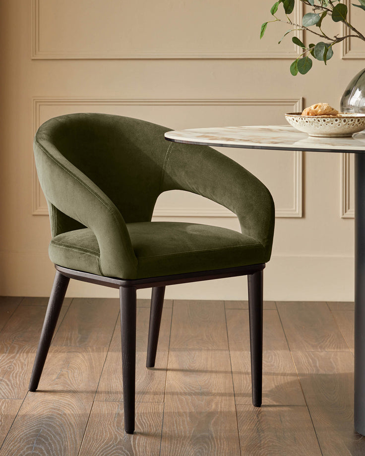 An olive velvet dining chair with dark wood legs sits next to a marble-topped table in a stylish dining space.