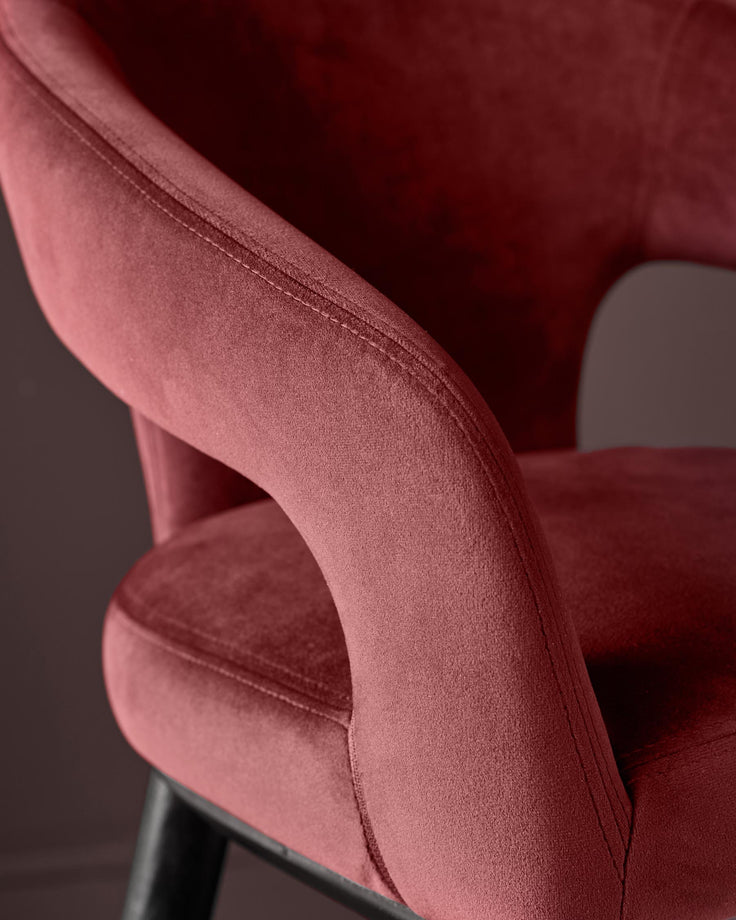 Close-up of a plush, velvet-upholstered chair in a deep maroon. Features curved armrests and dark wood legs.