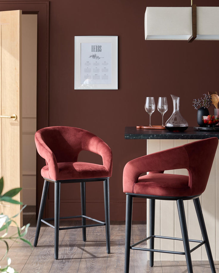 Stylish kitchen featuring two red velvet bar stools with black legs set around an island unit. Pictured in a warm lit setting.