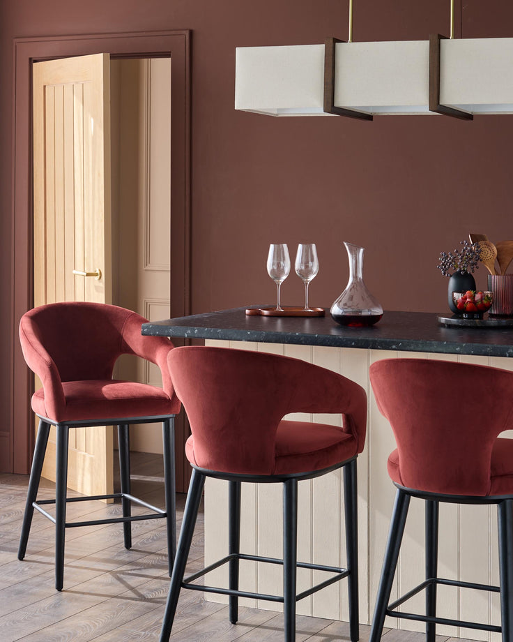 Three rust-coloured velvet bar stools sit at a kitchen island with a dark countertop, complemented by overhead lighting.