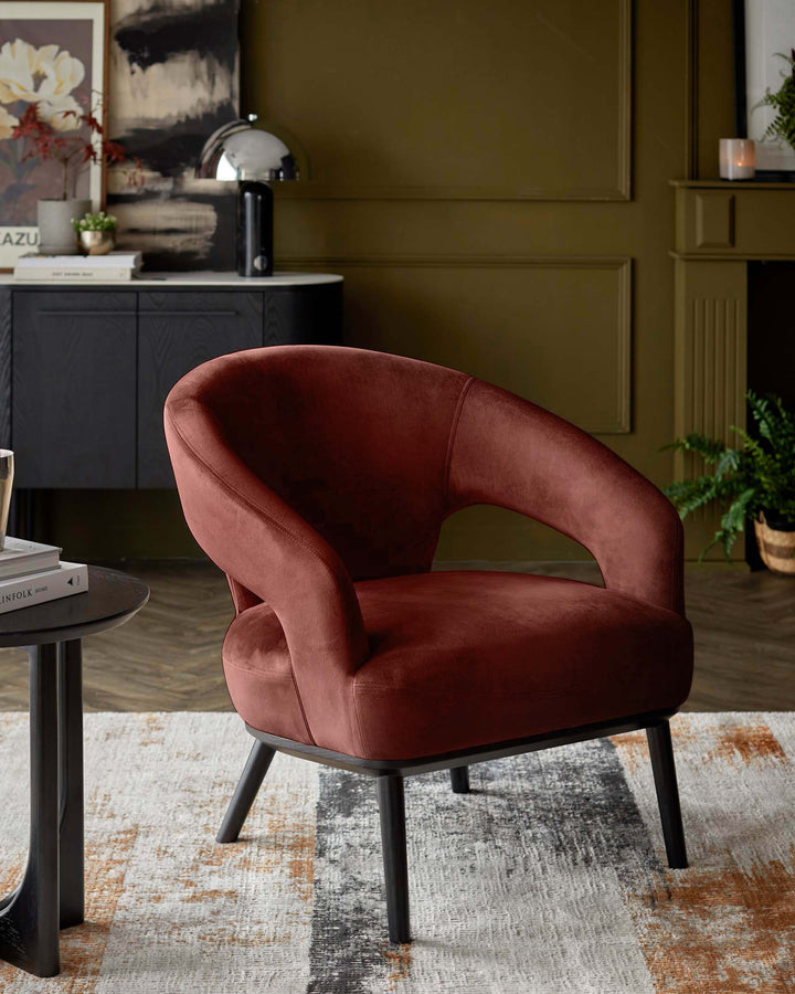 A stylish, rust-coloured velvet armchair with black legs sits on a patterned rug in a modern living room.