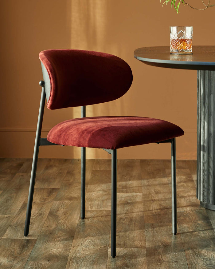 A velvet, auburn dining chair with a black metal frame sits next to a round table with a glass of whiskey on top.