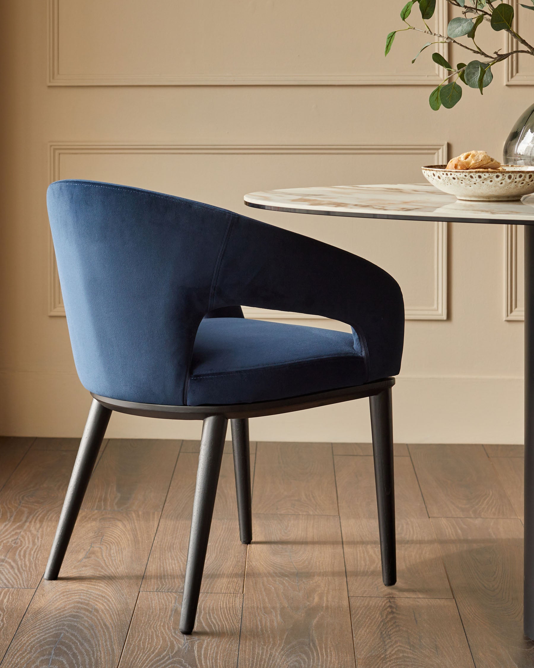 A stylish blue velvet dining chair with black legs sits next to a modern marble-topped table in an elegant dining setting.