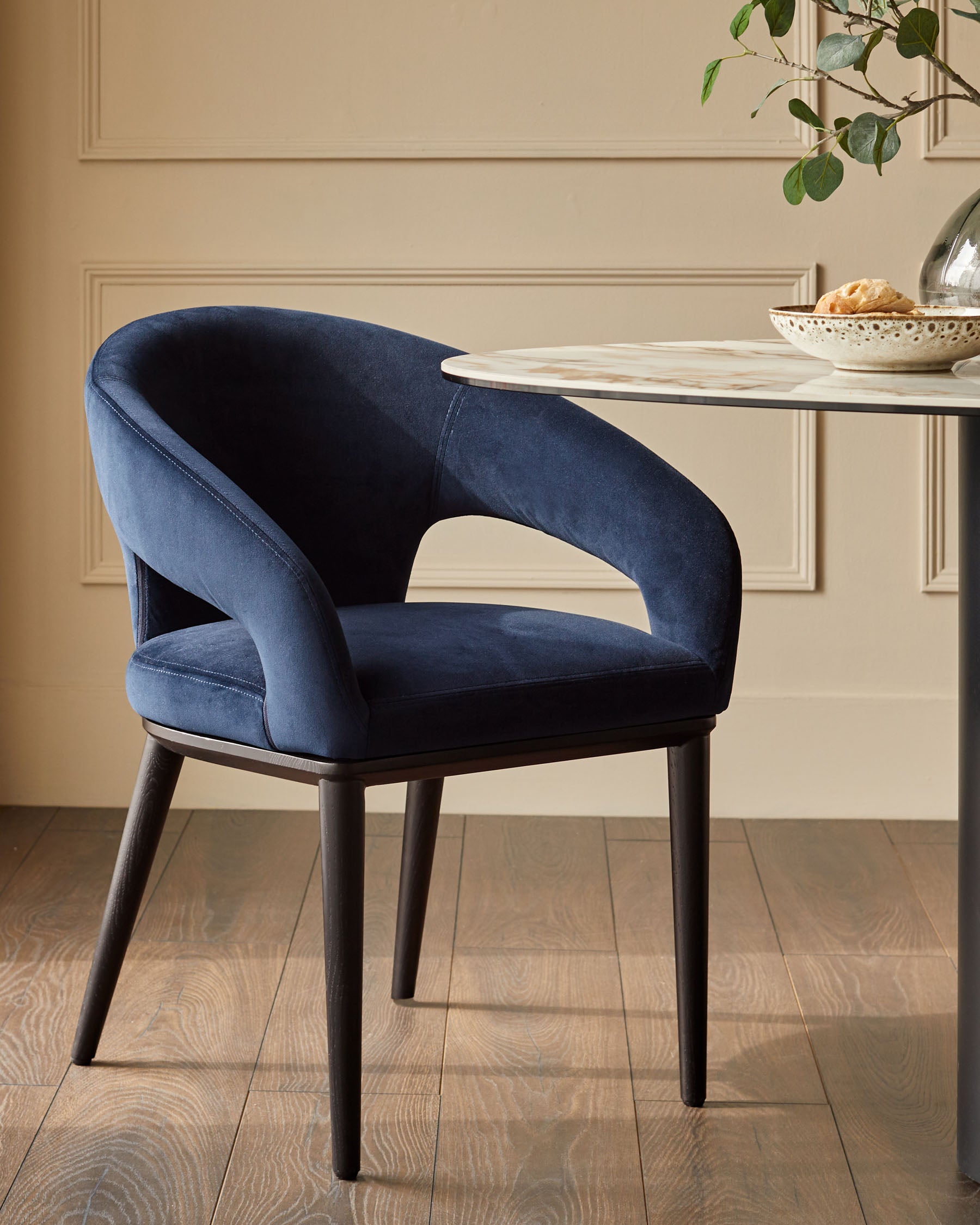 A luxurious blue velvet chair with a dark wooden frame sits beside a marble-topped table in a well-lit room.