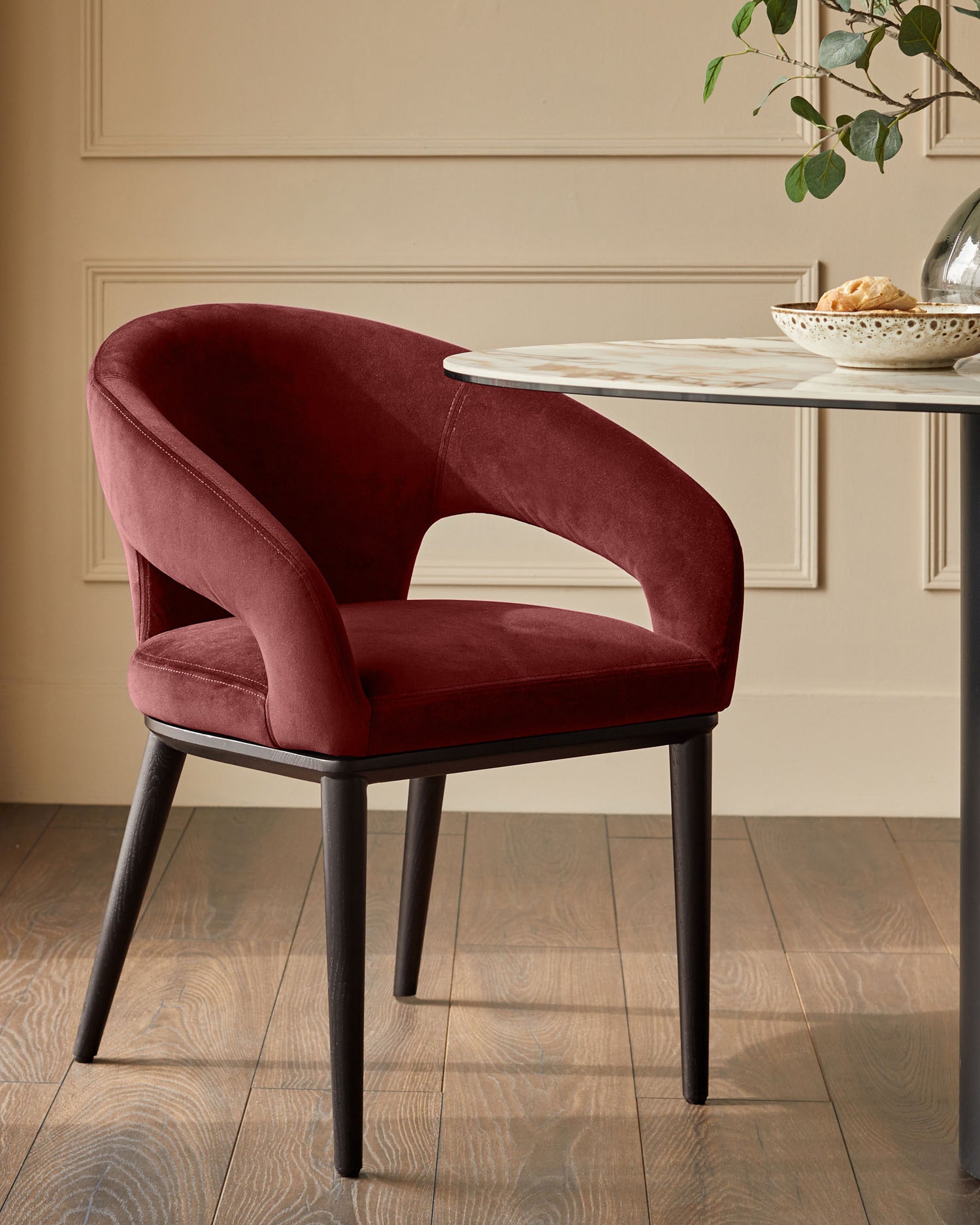 A stylish, contemporary dining chair in deep red velvet, with dark wooden legs, sits beside a round marble-topped table.