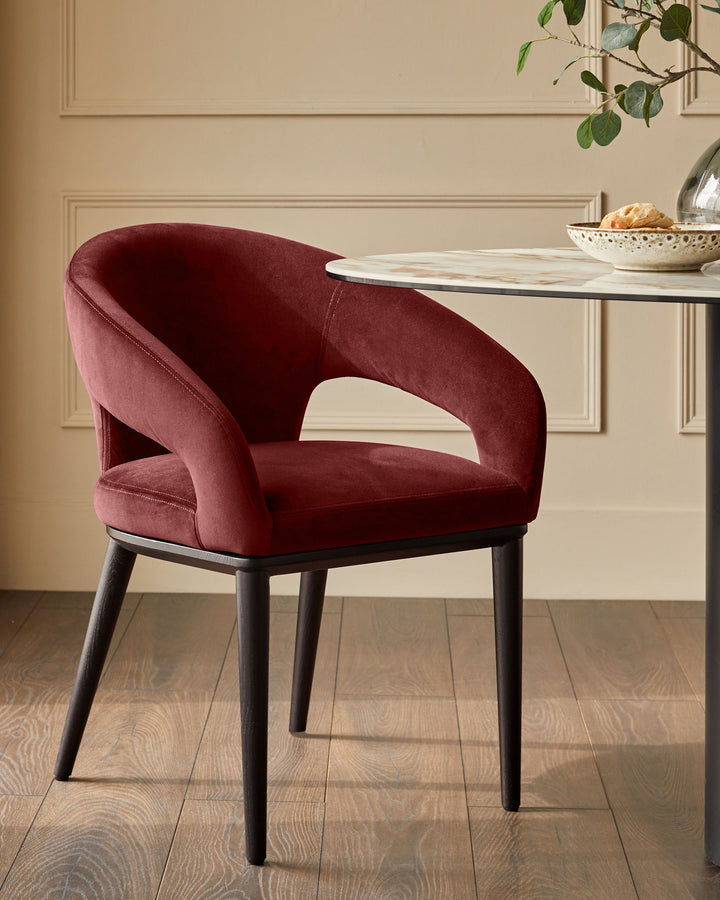 A stylish, contemporary dining chair in deep red velvet, with dark wooden legs, sits beside a round marble-topped table.