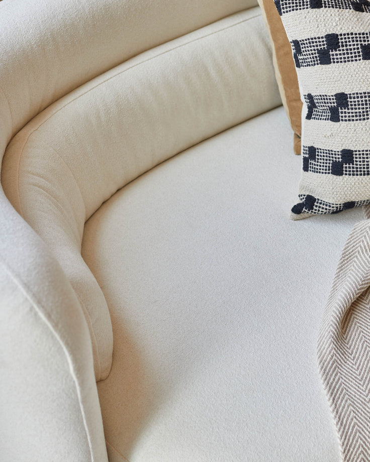 Close-up of a cream sofa, with a patterned cushion and throw. The sofa features a curved design.