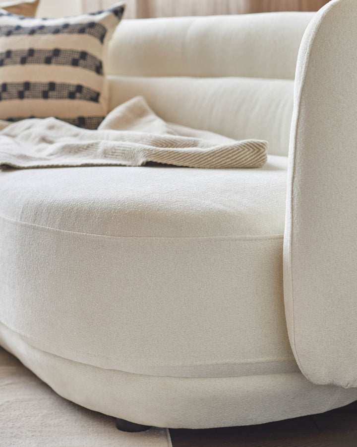 Close-up of a stylish, neutral-toned round sofa with a textured finish, accessorised with a patterned cushion and throw.