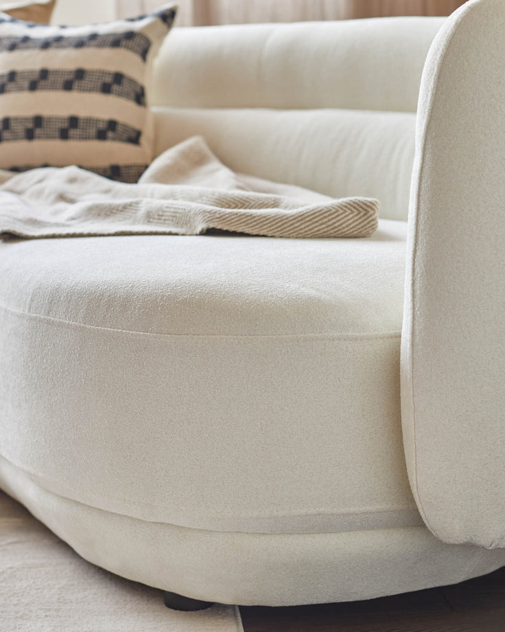 Close-up of a stylish, neutral-toned round sofa with a textured finish, accessorised with a patterned cushion and throw.
