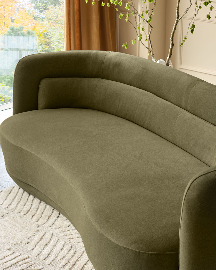 Henry 3-seater sofa in olive green velvet, curved design, positioned in bright living room with window and potted plant.