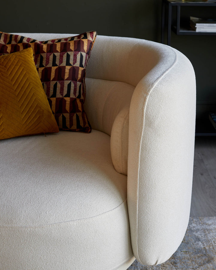 Cream curved sofa with mustard yellow and geometric patterned scatter cushions, with a dark shelf unit in the background.