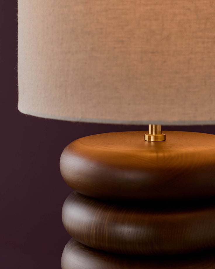 Hepworth table light with stacked dark wood base, brass connector, and natural linen shade against burgundy background.
