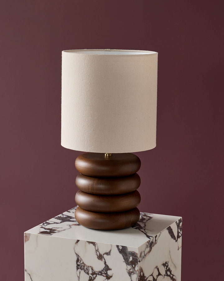Hepworth table lamp with stacked dark wood base and natural linen cylinder shade on marble-effect pedestal.