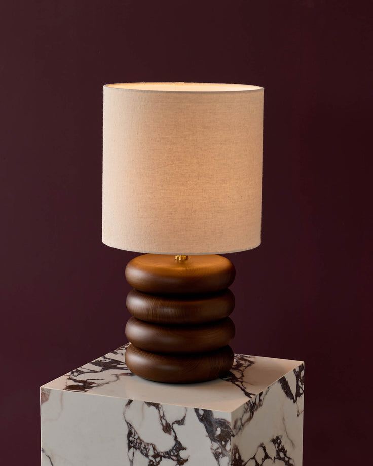 Hepworth table lamp with stacked dark wood base, natural linen shade, on marble-effect pedestal.
