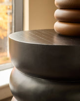 Black mango wood side table with stacked cylindrical base, shown at close-up angle near window
