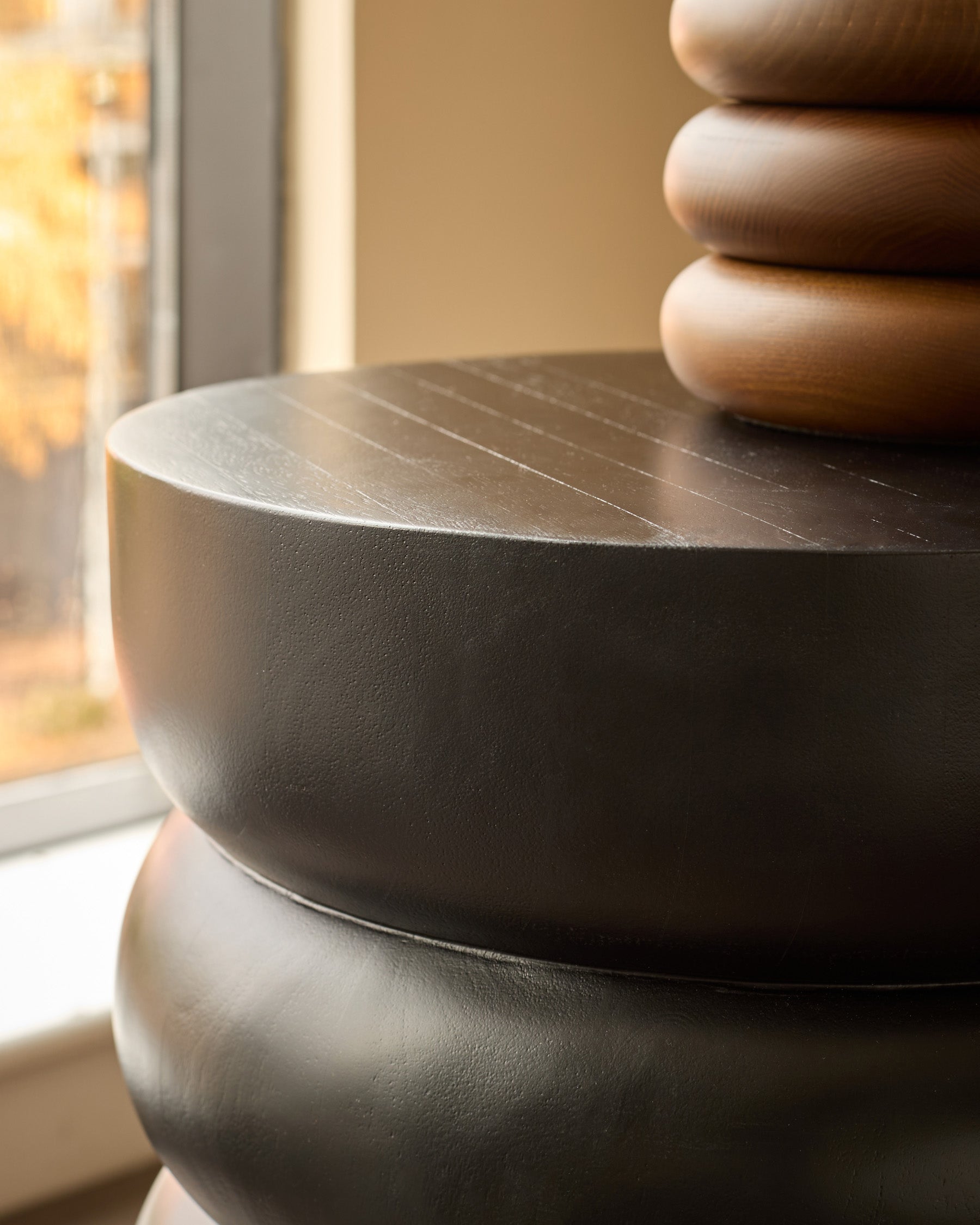 Black mango wood side table with stacked cylindrical base, shown at close-up angle near window