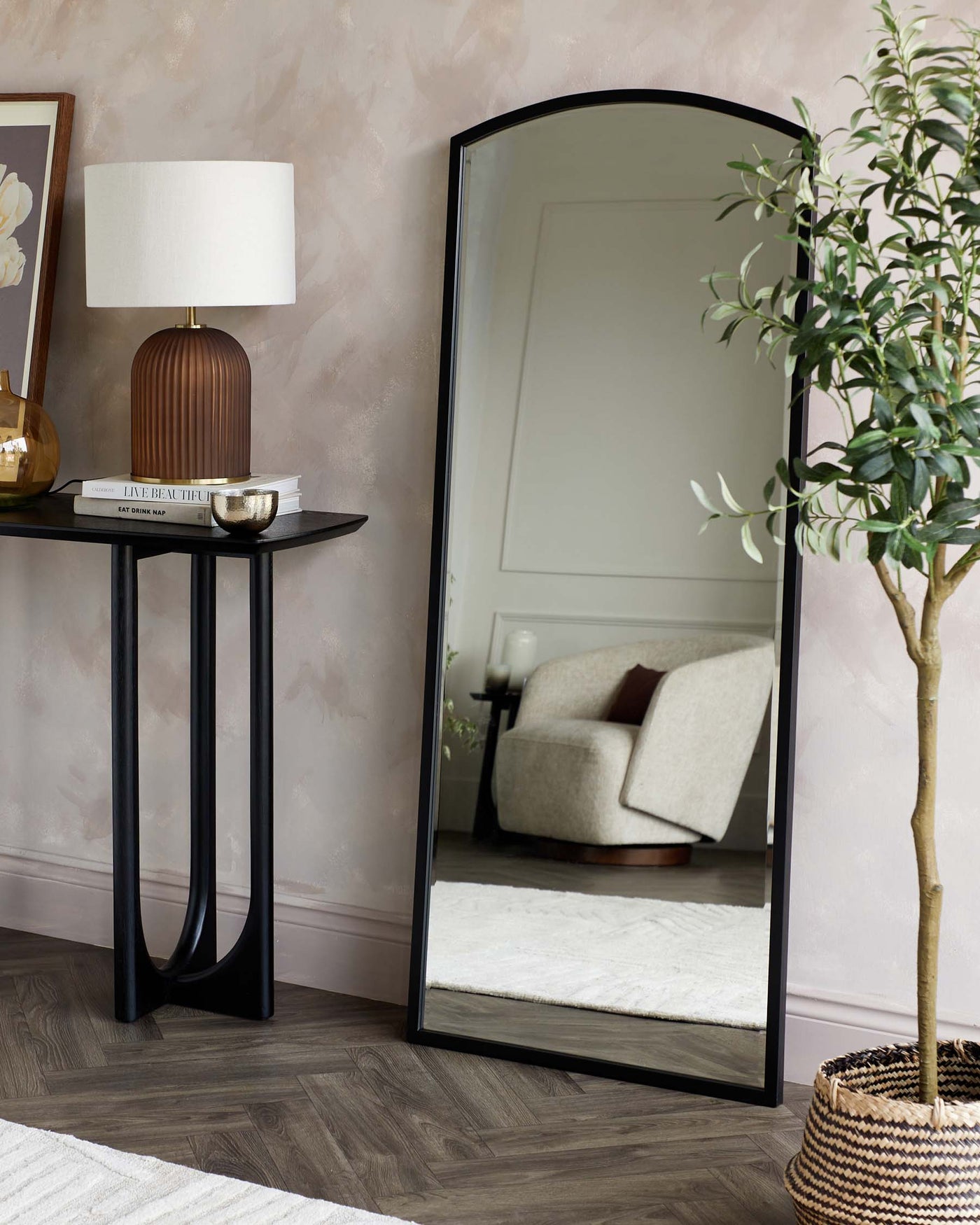 A sleek, modern black console table with a rectangular top and unique curved legs, paired with a large, arched floor mirror with a minimalist black frame. On the table rests a ribbed, cylindrical brown table lamp with a white shade, alongside a small stack of books and a decorative metallic bowl.