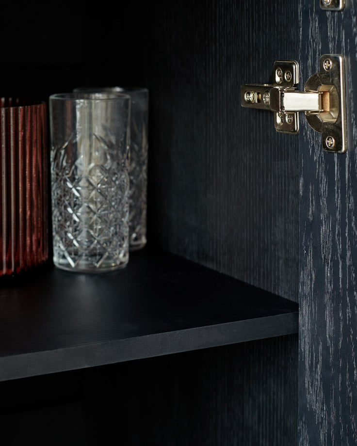 Close up on a modern black cabinet shelf with clear textured tumblers, and a hinge in view.