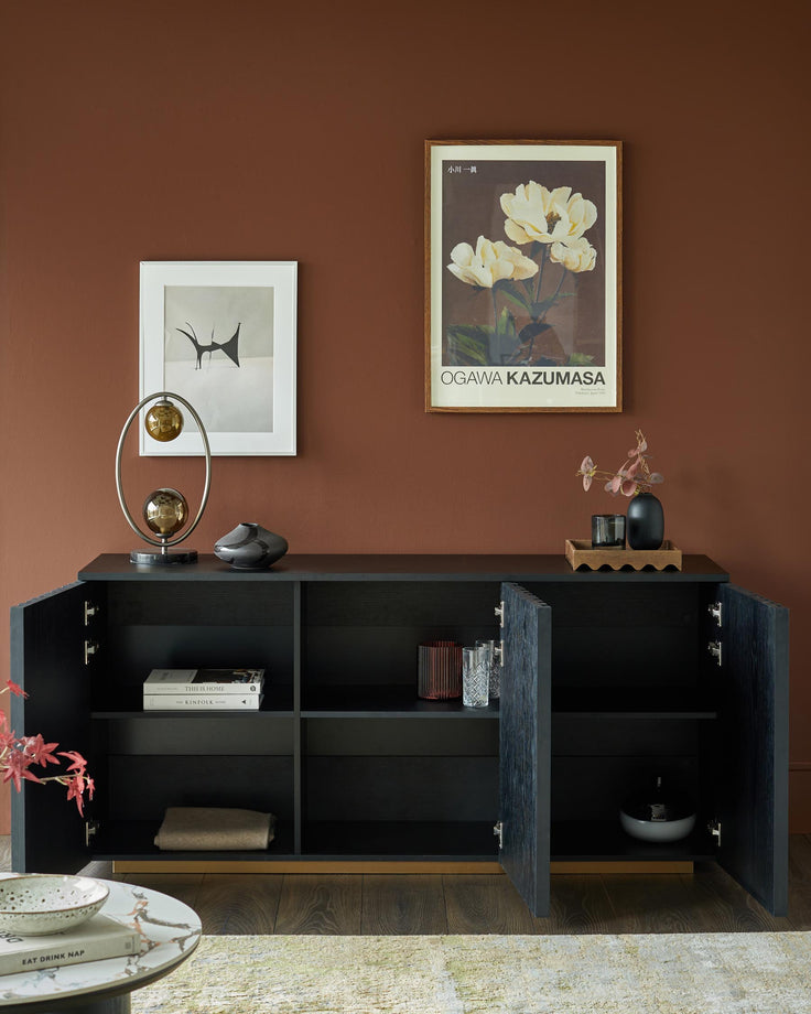 A dark wood sideboard with open shelving displays books, glassware and decorative items against a brown wall with framed prints.