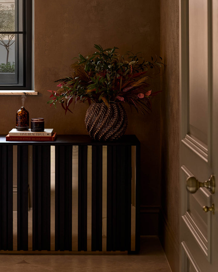 Holland sideboard in black with brass/gold vertical slats, styled with plants and books in a warm-toned interior setting.