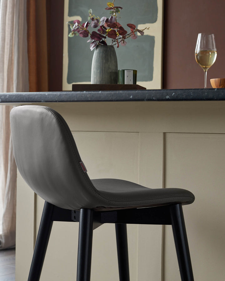 A modern bar stool with a grey leather seat and black legs sits next to a kitchen counter top in a stylish interior.