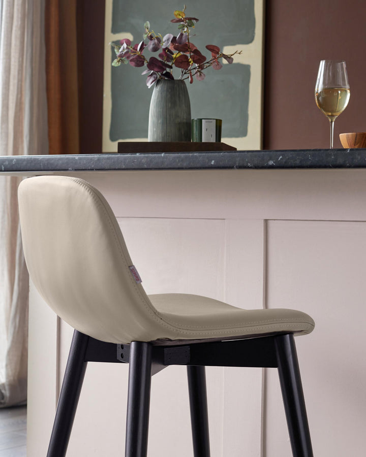 A light beige bar stool with dark wooden legs sits beneath a counter. A vase and wine glass are displayed behind.