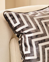 Imani cushion in chocolate with geometric striped pattern, displayed on cream upholstered furniture with leather piping detail.
