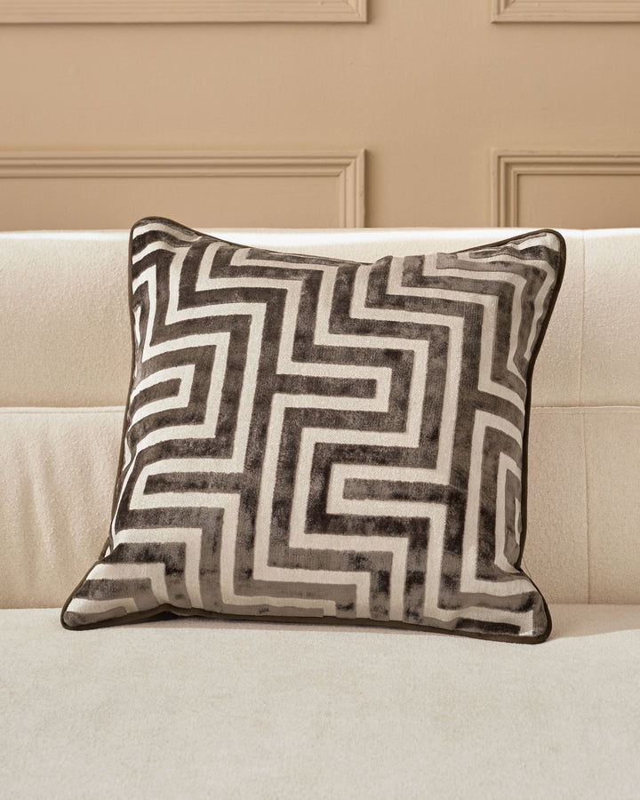 Imani cushion in chocolate with geometric maze pattern, displayed on cream sofa against beige paneled wall.