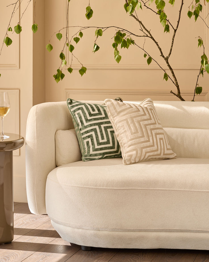 Imani cushion in olive green with geometric pattern, displayed on cream curved sofa in modern living room setting.