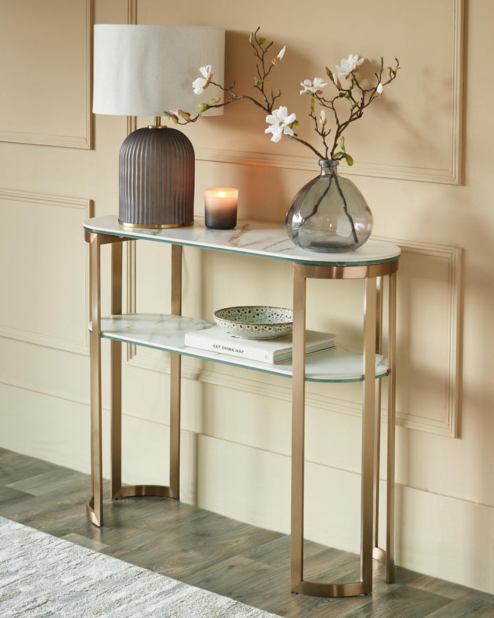 A modern console table with a marble effect top and glass shelf. Table lamp, candle, flowers and book are displayed on the table.