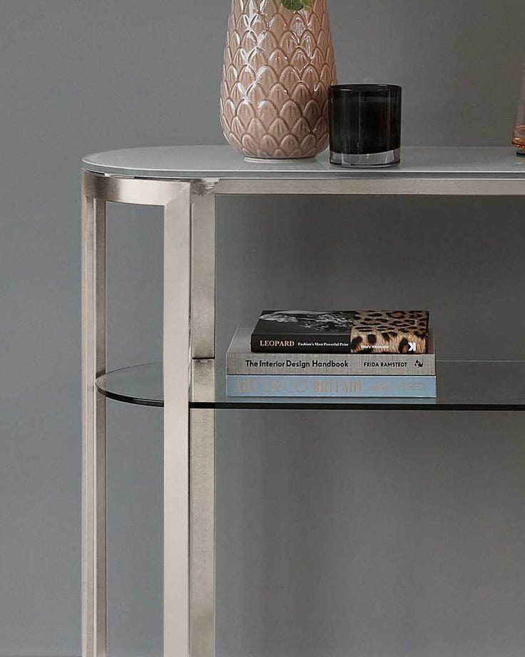 Isla console table in white with stainless steel frame, glass shelves, displaying books and decorative items on top.