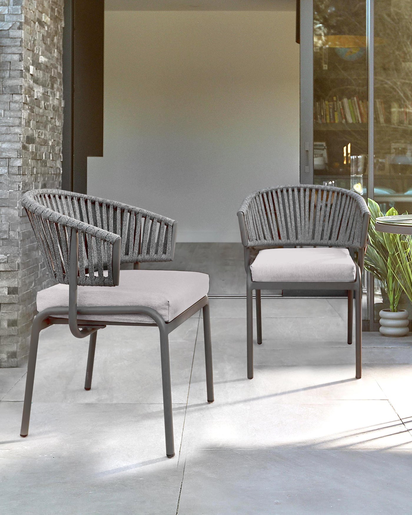 Two contemporary outdoor chairs with a minimalist design, featuring a matte metallic frame and vertical cordage backrest, complemented by smooth, light-coloured seat cushions. The chairs are set on an outdoor patio area, suggesting a modern and stylish outdoor living space.