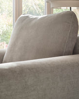 Jacob armchair in light grey fabric, shown at an angle displaying the padded backrest and armrest detail near a window.