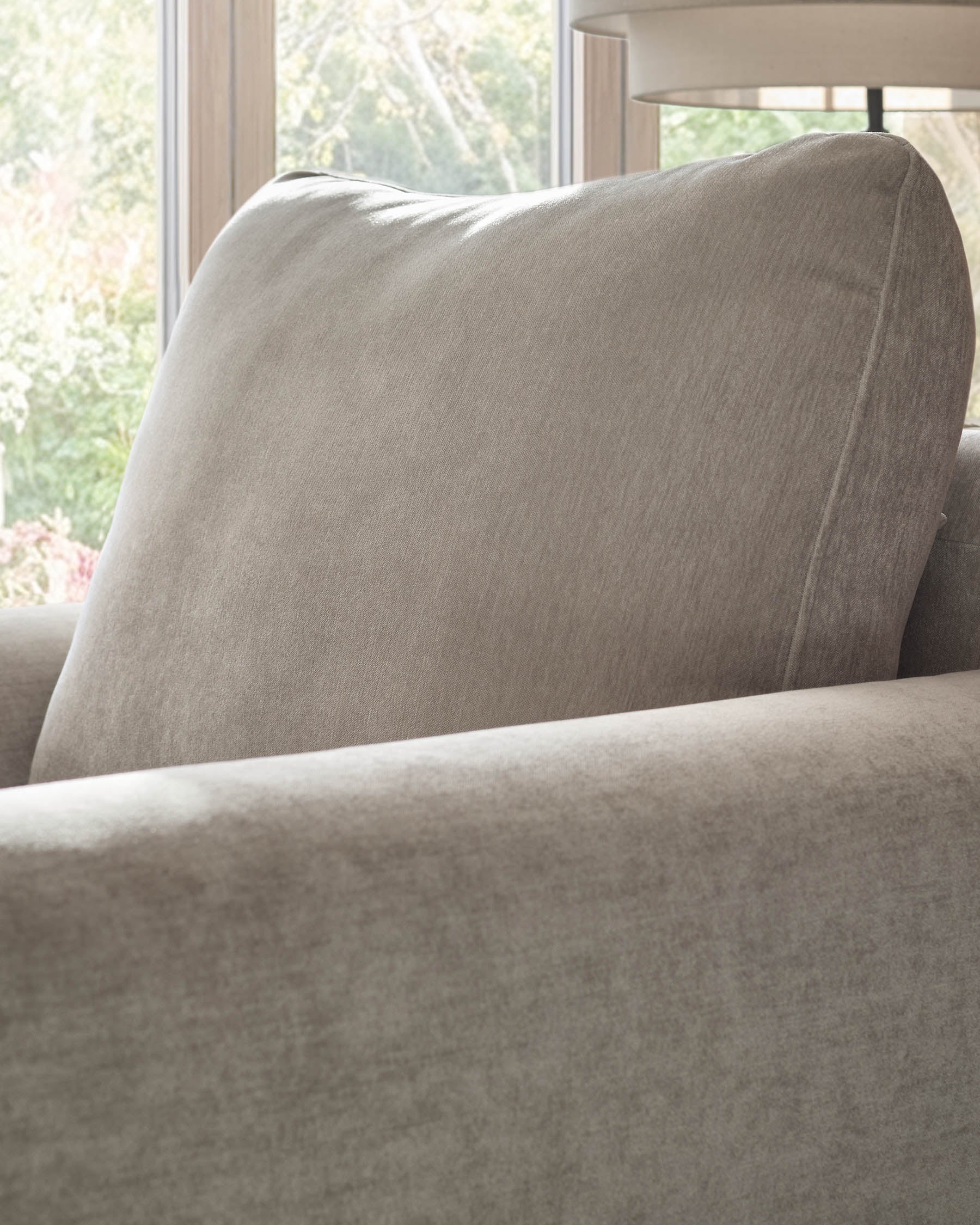 Jacob armchair in light grey fabric, shown at an angle displaying the padded backrest and armrest detail near a window.