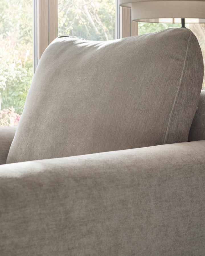 Jacob armchair in light grey fabric, shown at an angle displaying the padded backrest and armrest detail near a window.