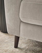 Light grey fabric armchair with curved arms, shown from side angle on textured beige floor with dark wooden leg visible.