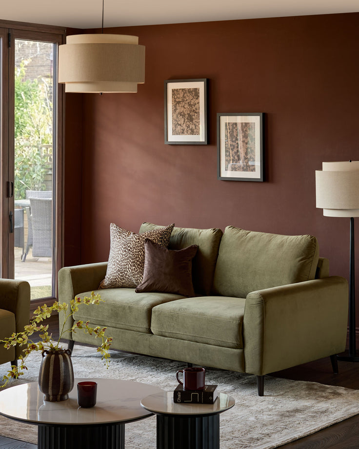 Jacob 3-seater sofa in olive green velvet, shown in a warm brown living room with decorative pillows and modern styling.