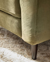Olive green velvet armchair leg detail on textured neutral rug, showing metal tapered leg and upholstered base corner.