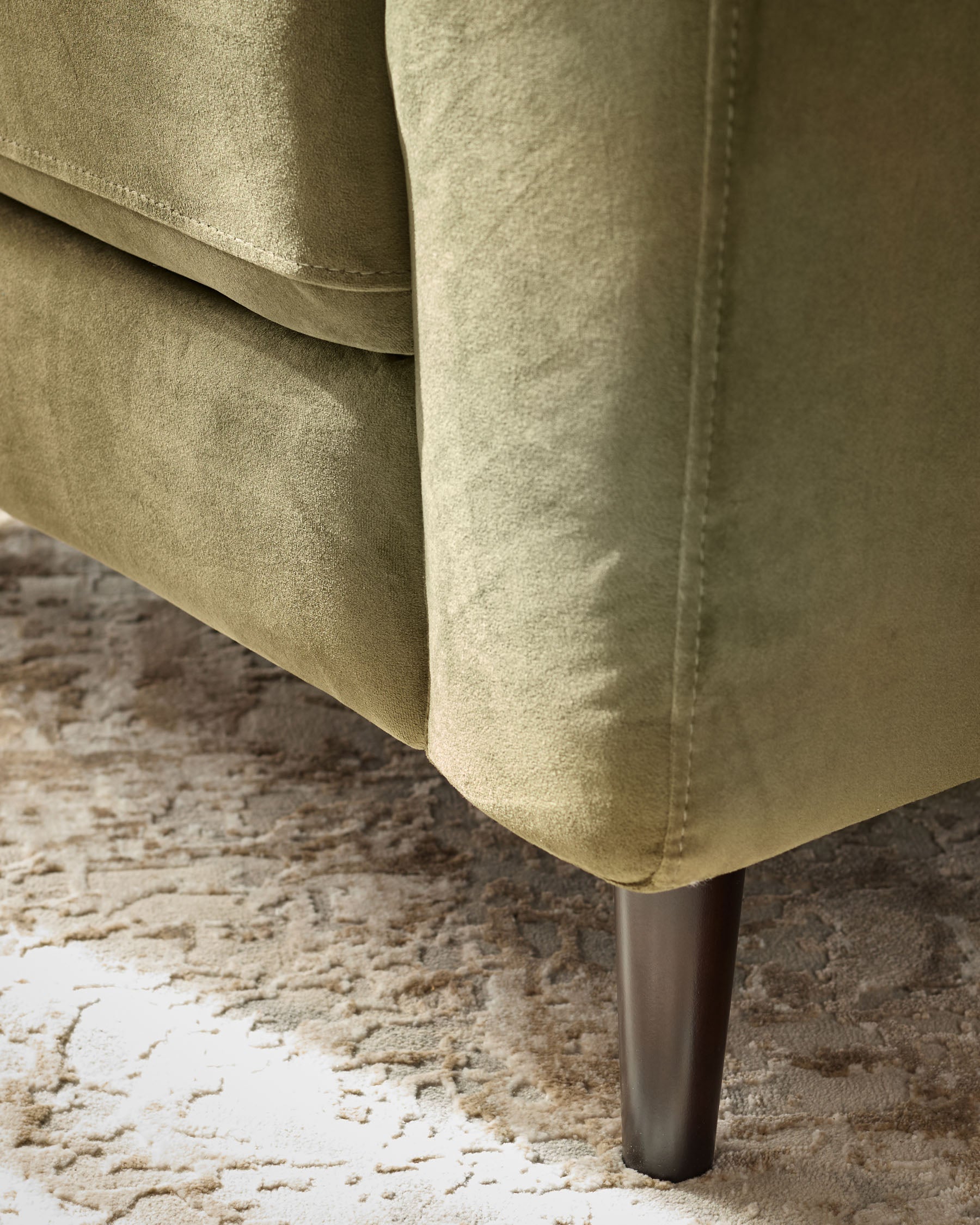 Olive green velvet armchair leg detail on textured neutral rug, showing metal tapered leg and upholstered base corner.