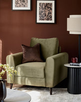 Jacob armchair in olive green velvet with dark wood legs, styled with a brown cushion against a burgundy wall.