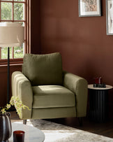Jacob armchair in olive green velvet, displayed in a styled living room with warm brown walls and natural lighting from windows.
