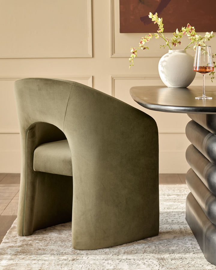 Jasper dining chair in olive green velvet, shown at an angle beside a round table with white vase and wine glass.
