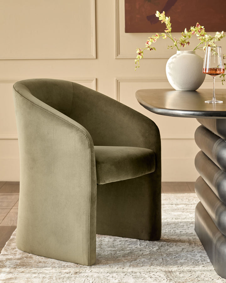 Jasper dining chair in olive green velvet, shown at an angle beside a round table with floral arrangement and wine glass.